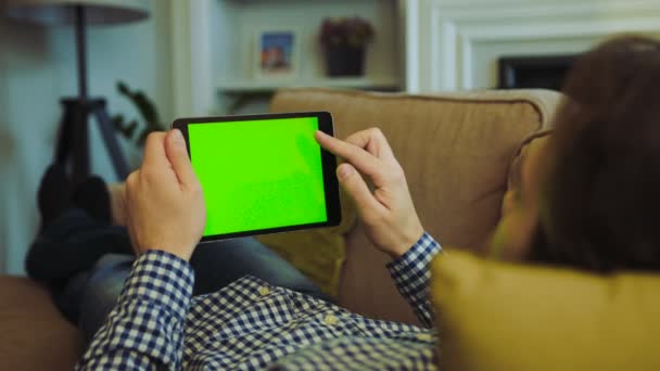 Blick über die Schulter auf den Mann in kunterbuntem Hemd und Jeans, der auf der Couch liegt und auf dem grünen Bildschirm eines schwarzen Tablet-Computers in seinen Händen blättert. Chroma-Schlüssel. Ruhe zu Hause. Innenräume — Stockvideo