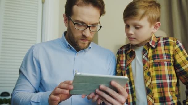 Pai de óculos usando seu dispositivo tablet e tendo problemas com ele para que seu filho jovem levá-lo para ajudar seu pai. Em casa. Interior — Vídeo de Stock