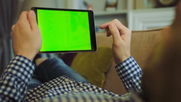Мужчина в клетчатой рубашке и джинсах лежит на уютном диване и прокручивает на зеленом экране черного планшетного устройства в руках. Хрома-ключ. Вид через плечо. Закрывай. Отдыхает дома. В помещении — стоковое видео