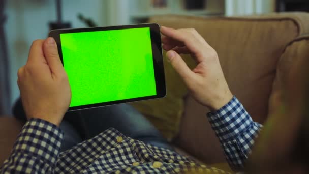 Vista sobre el hombro del hombre en camisa a cuadros y jeans acostados en el sofá con el dispositivo de tableta negro en las manos. Pantalla verde. Clave de croma. Descansando en casa. Adentro. — Vídeos de Stock