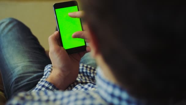 Rengarenk gömlek ve kanepede oturan ve onun elinde siyah smartphone yeşil ekranda kaydırma kot erkekte omzuna üzerinde görüntüleyin. Chroma anahtar. Yakın çekim. Kapalı — Stok video