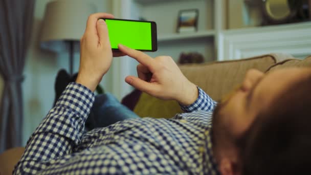 Hombre con la camisa a cuadros y jeans acostado en el sofá y desplazándose y grabando en la pantalla verde del teléfono inteligente negro horizontal en sus manos. Clave de croma. Tiempo de descanso en una habitación acogedora. De cerca. De interior — Vídeo de stock