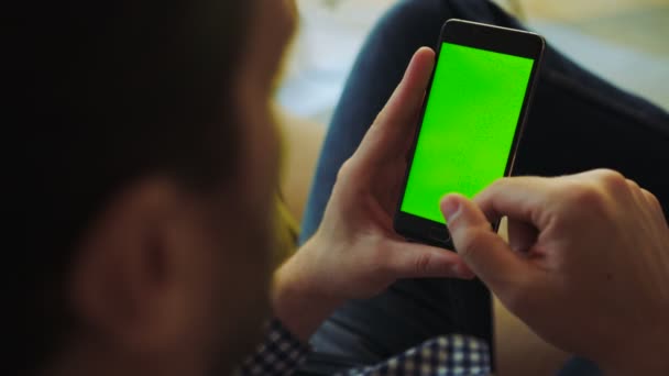 Ver por encima del hombro en el hombre en la camisa a cuadros y vaqueros sentados en el sofá y desplazándose en el teléfono inteligente negro en sus manos. Clave de croma. Pantalla verde. De cerca. Adentro. — Vídeos de Stock