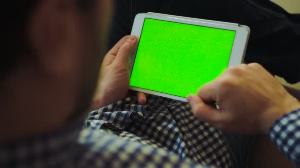 Vue au-dessus de l'épaule sur l'homme en chemise bigarrée et en jeans assis sur le canapé et faisant défiler sur l'écran vert de la tablette blanche dans ses mains. Clé chromatique. Ferme là. Intérieur — Video