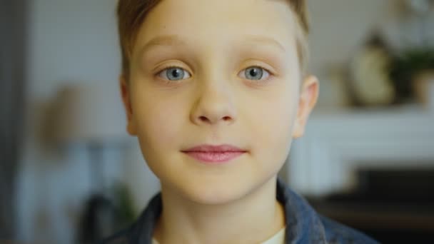 Close up do menino bonito loiro com olhos azuis em pé na frente da câmera na sala de estar, olhando em linha reta e sorrindo. No interior. Retrato tirado — Vídeo de Stock