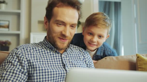 Närbild på den Söt blond liten pojke tittar på sin far som gör något på den bärbara datorn, både ler och pratar. Hemma på soffan. Inuti — Stockvideo