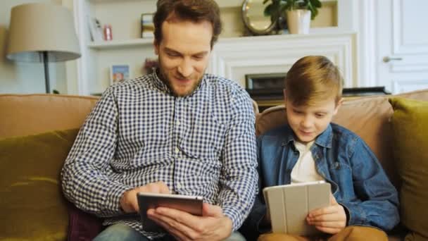 Portrét shot z otce a syna, jak sedí na gauči v obývacím pokoji, mluví a používání jejich zařízení tablet. Doma. Uvnitř — Stock video