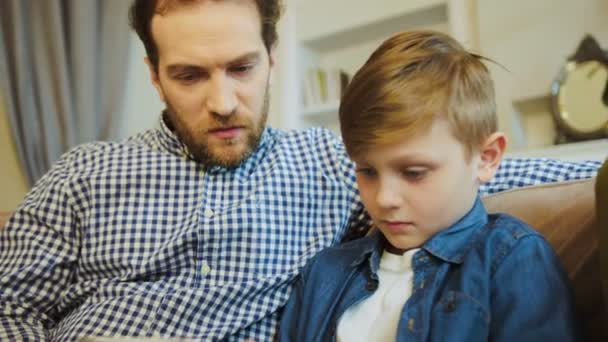 Primer plano del padre en una camisa abigarrada mirando a su pequeño hijo encantador sosteniendo la computadora de la tableta y jugando en ella. En casa, en el sofá. De interior — Vídeo de stock