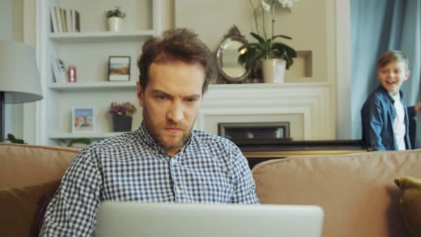 Portrait du beau père dans une chemise à carreaux et avec barbe travaillant sérieusement sur l'ordinateur portable sur le canapé à la maison. Petit fils futé qui vient à lui par la porte arrière. Intérieur — Video