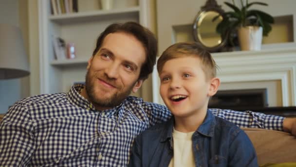 Primer plano del padre guapo con una barba y un niño lindo, su hijo, viendo la televisión en el sofá en la sala de estar y riendo. Un retrato. De interior — Vídeos de Stock