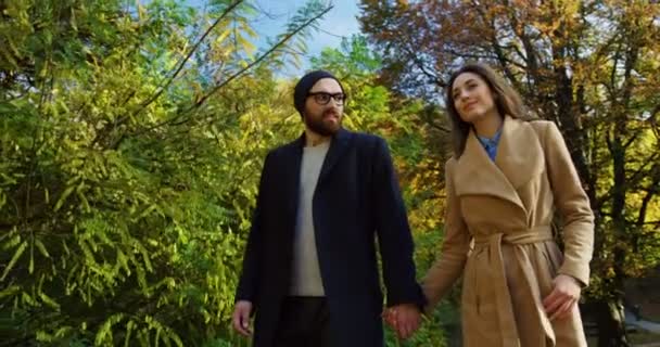 Щаслива усміхнена пара в пальто, що йде рукою в руці в парку на початку осені. Сонячний день. На відкритому повітрі — стокове відео