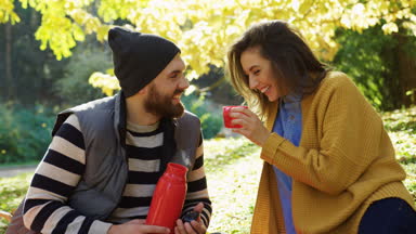 Bir termos sıcak çay ile parkta oturan, konuşan ve gülen aşık genç çift portresi. Sonbaharda güneşli bir gün. Açık havada