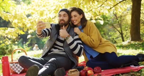 junge schöne Paar Video-Chat, zeigt Daumen hoch und lacht auf dem Smartphone in der gelben schönen Park an einem sonnigen Tag im Herbst. im Freien