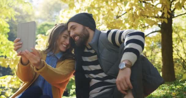 Retrato de una joven pareja romántica tomando hermosas selfies en el dispositivo de tableta entre los árboles amarillos en el parque. Soleado hermoso día de otoño. Exterior — Vídeo de stock