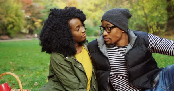 Retrato de una joven pareja afroamericana atractiva sentada en el picnic, mirándose unos a otros y que sonriendo directamente a la cámara. Día soleado en el parque de otoño. Exterior — Vídeos de Stock