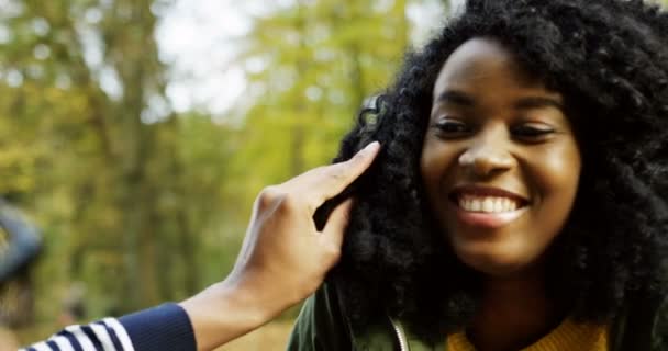 Крупным планом молодая симпатичная афроамериканка улыбается, а ее бойфренды играют с ее вьющимися волосами в парке. Хороший осенний день. На улице. Портрет — стоковое видео