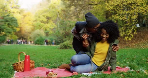 Jonge vrolijke Afro-Amerikaanse man en vrouw zittend op de rode deken op het gras, het kussen en het plezier in het park in de zonnige herfst. Buitenshuis — Stockvideo