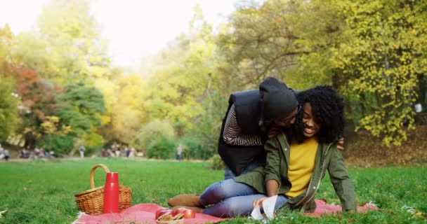 Jovem casal afro-americano feliz apaixonado sentado no cobertor vermelho na grama, beijando e abraçando. Estacione no dia ensolarado do outono. Ao ar livre . — Vídeo de Stock