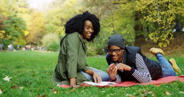 非洲裔美国人夫妇在公园里度过他们的时间很好。年轻女子坐在一本书和年轻男子躺在她旁边的智能手机。秋天晴朗的日子。外面 — 图库视频影像
