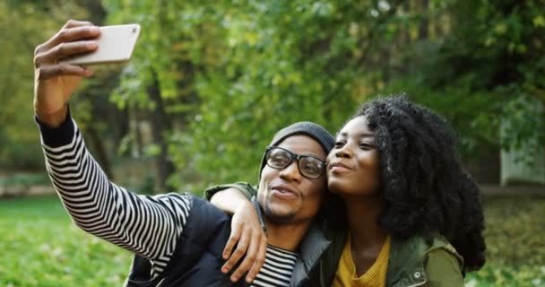 Junges attraktives afrikanisch-amerikanisches Paar, das an einem sonnigen Tag im herbstlichen Park Selfies mit dem Smartphone macht. Außenbereich — Stockvideo