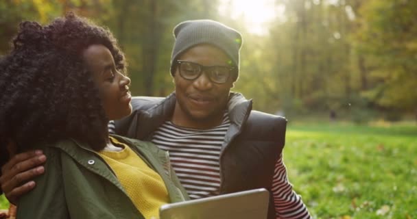 Retrato de jovem casal romântico afro-americano sentado perto um do outro, abraçando e olhando para a tela do computador tablet. Estacione no outono ensolarado. Ao ar livre — Vídeo de Stock