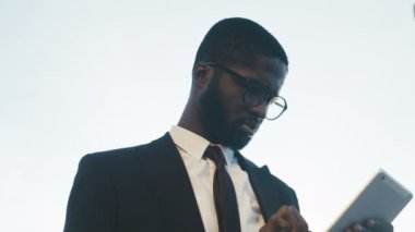 Bir kravat ve onun tablet bilgisayarda dokunarak ve gülümseyen Gözlük Genç yakışıklı Afrikalı-Amerikalı bir işadamı kapatın. Beyaz gökyüzü arka plan. Açık havada. Portre vurdu