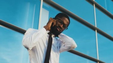 Genç Afro-Amerikalı işadamı kravat ve akıllı telefonda konuşurken ve office windows arka plan boynuna kadar ısınma gözlük bıktınız. Sorunlarınız ve endişe erkek ofis çalışanı