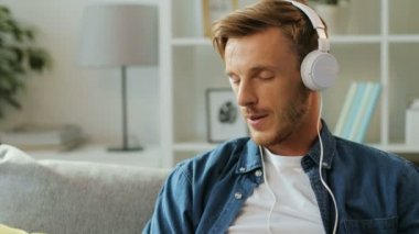 Akıllı telefon üzerinde beyaz kulaklık müzik dinlemek ve beyaz zemin üzerine evde kanepede otururken ritim başını sallayarak şık genç adam.