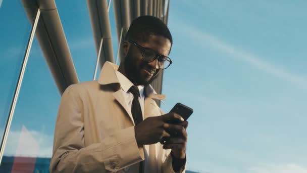 Крупным планом снизу на молодого привлекательного афроамериканского бизнесмена в галстуке и очках, стучащего по смартфону и улыбающегося. Голубое солнечное небо и большой современный фон офисного здания. На открытом воздухе — стоковое видео