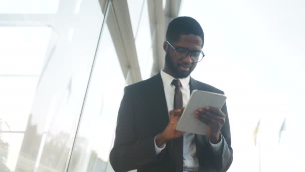 Ung snygg log African American affärsman i slips och glasögon trycka på sin tablett dator nära office centrum. Vit himmel bakgrund. Utomhus — Stockvideo