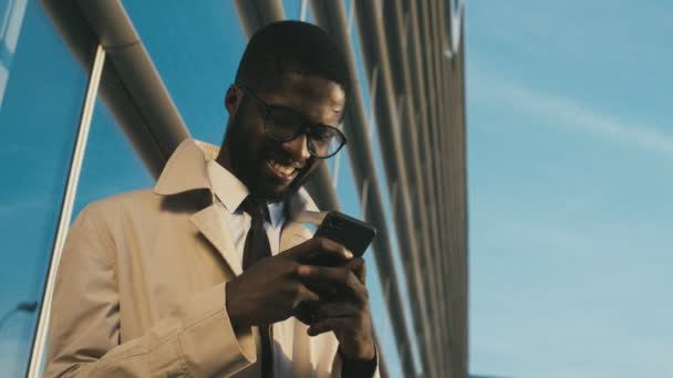 Ritratto dal basso sul giovane bell'uomo afroamericano che indossa cravatta e occhiali toccando il suo smartphone, sorridendo e chiudendo la bocca con una mano a causa di chiedersi. Cielo blu e un grande — Video Stock