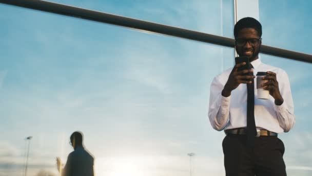 Молодой привлекательный афро-американский бизнесмен в галстуке, записанном на смартфон и пьющий кофе возле современного офисного центра в солнечный день. На открытом воздухе — стоковое видео