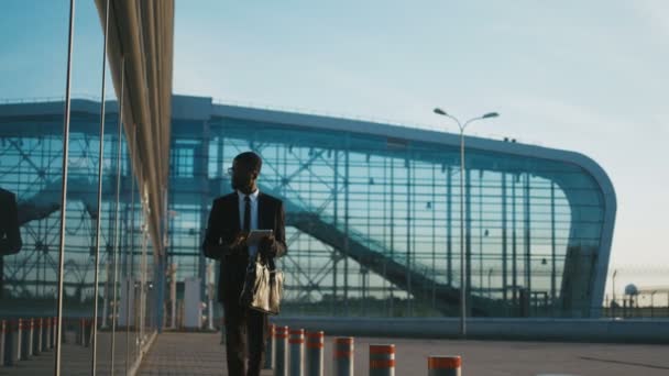 Bonito jovem empresário afro-americano de terno e gravata andando ao longo de um edifício do aeroporto e tocando no computador tablet. Fora do grande centro de vidro moderno . — Vídeo de Stock