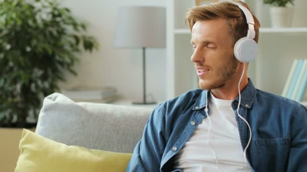 Junger stilvoller Mann hört Musik in den großen weißen Kopfhörern auf dem Smartphone, während er zu Hause auf dem Sofa auf weißem Hintergrund sitzt. — Stockvideo