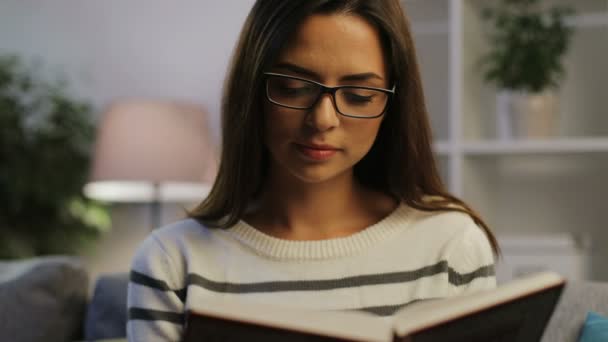 Retrato de una atractiva mujer joven relajándose en la elegante sala de estar y leyendo un libro interesante por la noche . — Vídeos de Stock