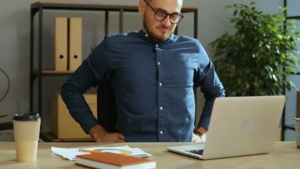 Homem de negócios atraente em camisa azul sentindo dor nas costas horrível, mas continuar trabalhando no laptop no escritório . — Vídeo de Stock