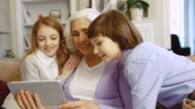 Büyükanne ile torunlarını evde kanepede otururken video sohbet için tablet kullanarak portresi.