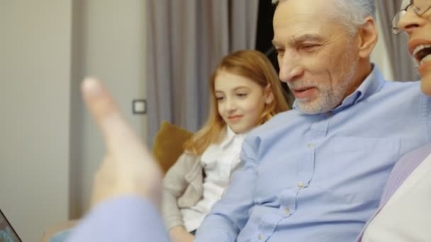 Vista lateral de los abuelos y nietos utilizando el ordenador portátil para el chat de vídeo mientras se sienta en el sofá en casa. Retrato plano . — Vídeos de Stock