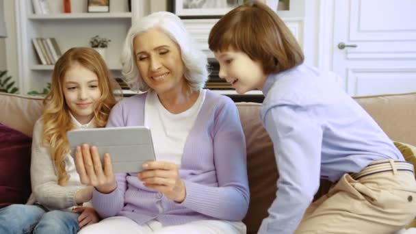 Hermosa abuela con sus nietos utilizando la tableta para el chat de vídeo mientras se sienta en el sofá en casa en la sala de estar . — Vídeo de stock