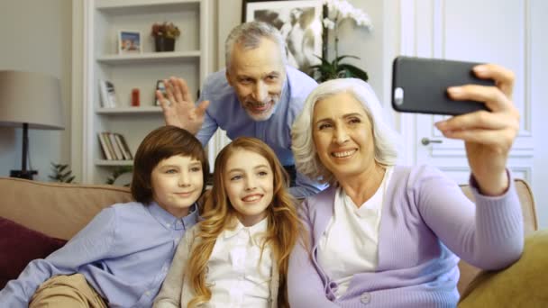 Portret van Kaukasische en gelukkige familie maken selfie met behulp van slimme telefoon zittend thuis in de woonkamer. Grootouders met kleinkinderen. — Stockvideo