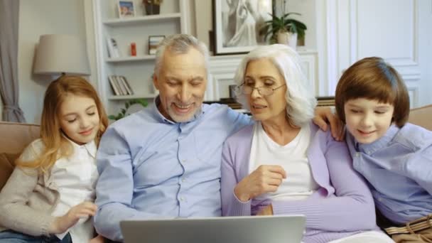 Heureux grands-parents et petits-enfants utilisant un ordinateur portable pour bavarder en vidéo assis sur le canapé dans le salon . — Video