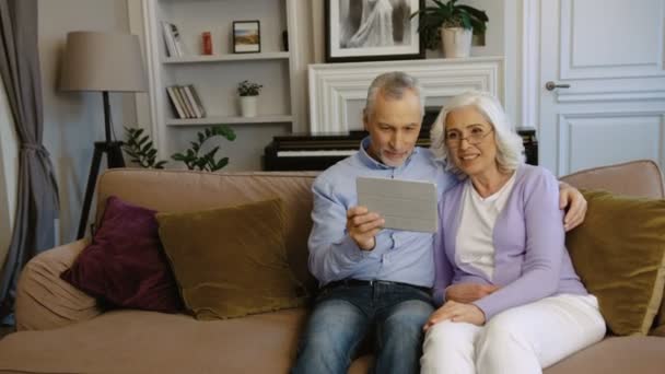 Sevimli büyükanne ve Büyükbaba evde şık oturma odasında kanepede otururken tablet kullanma. Yan görünüm. — Stok video