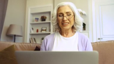 Mutlu yaşlı kadın evde oturma odasında sohbet video için dizüstü bilgisayar kullanarak bardaklarda görüntüsünü kapatmak.