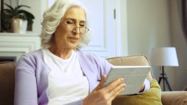 Güzel yaşlı kadın kanepe oturma odasında yorgunluk internette sörf için tablet kullanarak bardaklarda yan dikey görünüm.