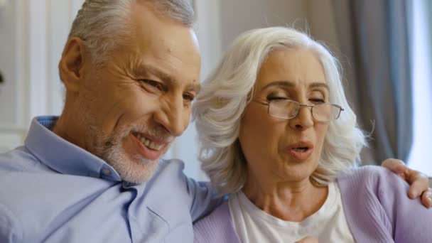 Primer plano de pareja de ancianos sentados en el sofá en casa y posando en la cámara al hacer selfie . — Vídeos de Stock