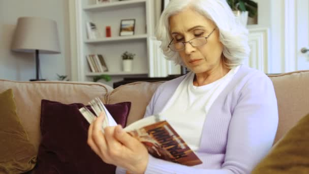 Portré vonzó idős asszony, kanapéval, elegáns otthon ül, és könyvet olvas. Beltéri lövés. — Stock videók