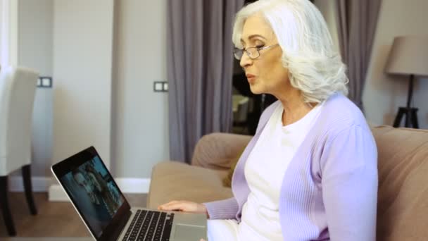 Retrato de anciana feliz en gafas usando el ordenador portátil para el chat de vídeo en casa en la sala de estar. Vista lateral . — Vídeos de Stock