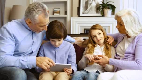 Портрет бабушки и дедушки с внуками с помощью планшета и разговоров во время отдыха на диване в гостиной . — стоковое видео