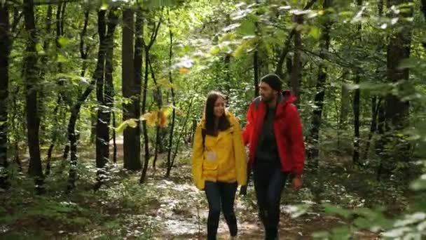 Jovem atraente mulher feliz e homem caminhando na floresta com mochilas e em casacos coloridos. Ao ar livre — Vídeo de Stock