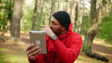 Onun tablet cihaza güzel güneşli ormandaki sohbet ve orman kameraya gösteren genç yakışıklı erkek uzun yürüyüşe çıkan kimse video portresi. Açık havada